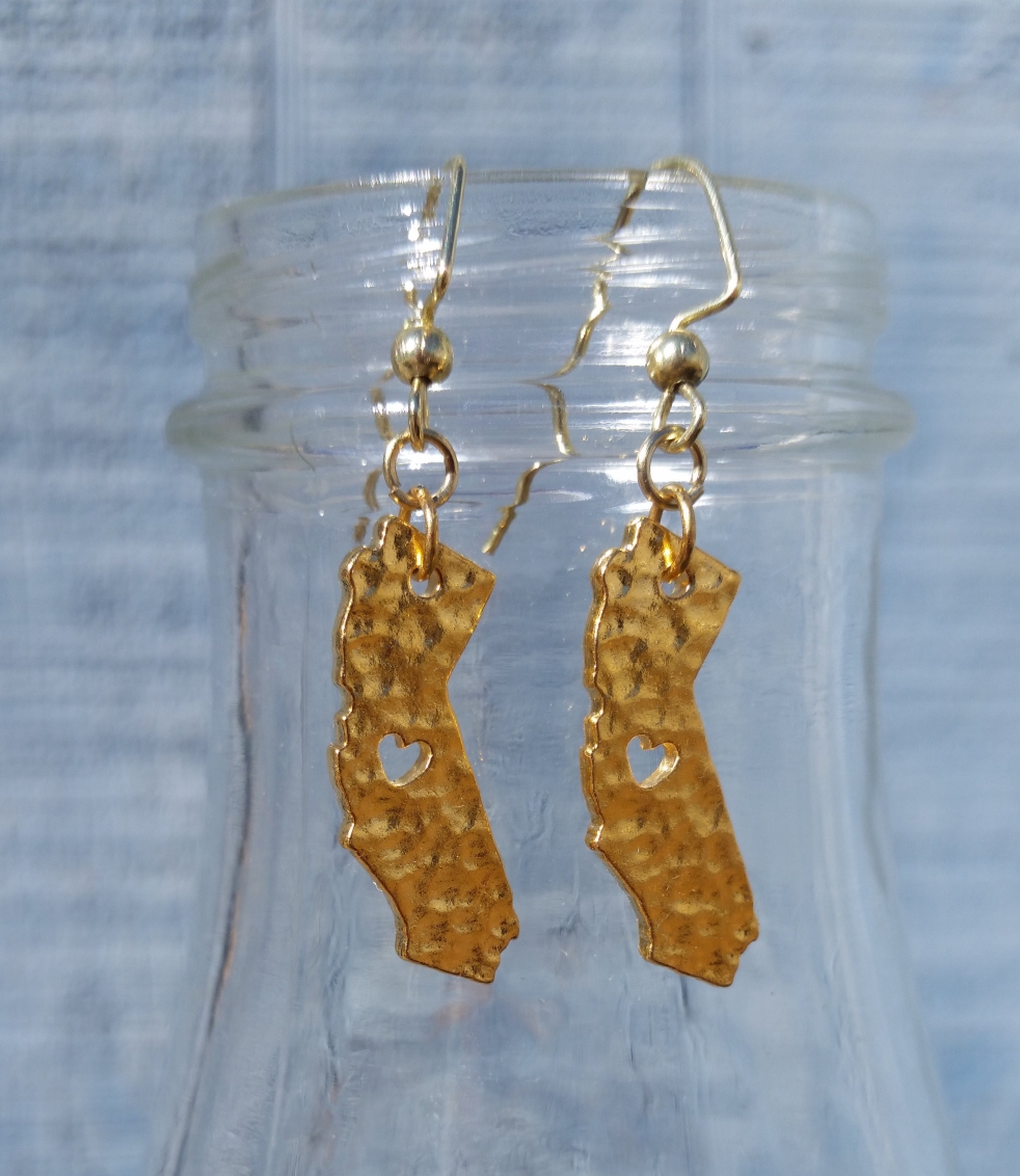 A pair of gold plated dangle earrings. Each earring has a charm with the state of California dangling from it.