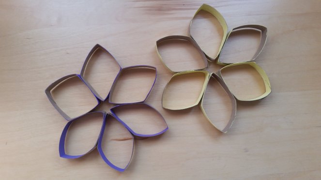 There are two snowflakes made from toilet paper rolls. There is a purple one and yellow one. Both have six points, thus, using six pieces glued side by side until they come full circle. Each component is in the shape of a diamond or parallelogram. Both the purple and yellow have three painted on the outside and three painted on the inside of each parallelogram. This photo shows both are laying flat on a table.