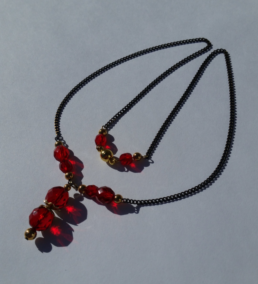 Necklace is laid in the shape like the white flower in the background/certain grassy stages of Super Mario games. There are 8 red beads and 10 gold beads, 18 in total. The pendant has 2 gold beads and 2 large red beads strung vertically. The pendant is connected to the front of the necklace with 4 red and 4 gold beads, two of each on either side, connected to black chain with gold shimmer. Chain on both sides are then connected to the back of the necklace where there is a round magnetic clasp surrounded by 2 gold beads and 1 red bead on either side.