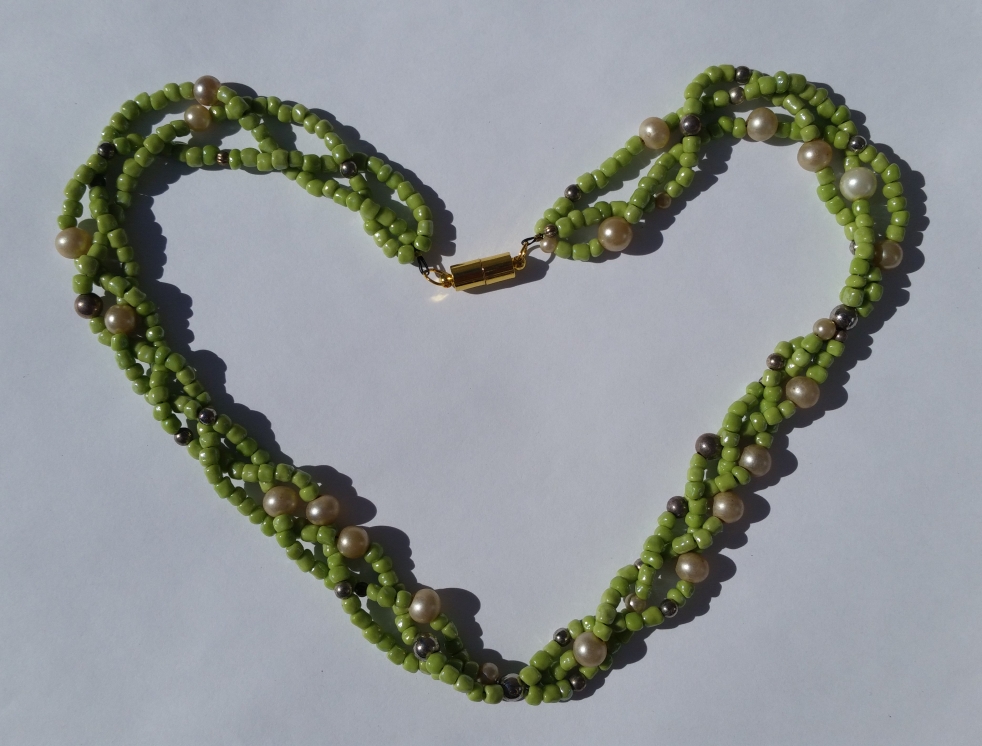 3 strands of beads are braided. Each strand has lime green, cream, and metal beads of different sizes. Each strand also has their own pattern. The necklace is closed with a tube-shaped magnetic clasp. Necklace is laid in the shape of a heart.